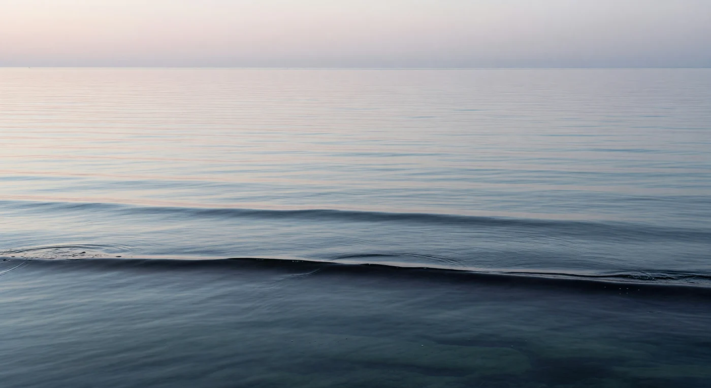 At the edge of night and day, the ocean surface lies almost perfectly still — a mer d'huile stretched beneath a brightening sky, where the air-sea interface is measured not in fathoms but in micrometers, a skin of water so thin it exists as chemistry more than depth. Dawn light arrives at an extreme angle, skimming the near-glassy plane and igniting cold pink and pearl reflections across the long dark swells that the submerged basalt ridge organizes from below, its volcanic geometry pressing its linear signature up through the water column as subtle banding visible only under such raking illumination. Within the sea-surface microlayer — that uppermost millimeter of ocean, biochemically distinct from everything beneath — dissolved organic films, lipid sheets, and microscopic life accumulate in a world of extraordinary concentration, where surface tension itself becomes an ecological force and neuston organisms navigate an interface between two atmospheres. The basalt ridge below, born of ancient seafloor spreading, slows and refracts the long swells into the faintest capillary corrugation, its dark flanks softened by refraction and traced in caustic shimmer as first light penetrates the upper meters with exceptional clarity. This is the ocean as it almost always is — vast, unhurried, indifferent, organizing itself according to physics and geology and biology with no witness required.