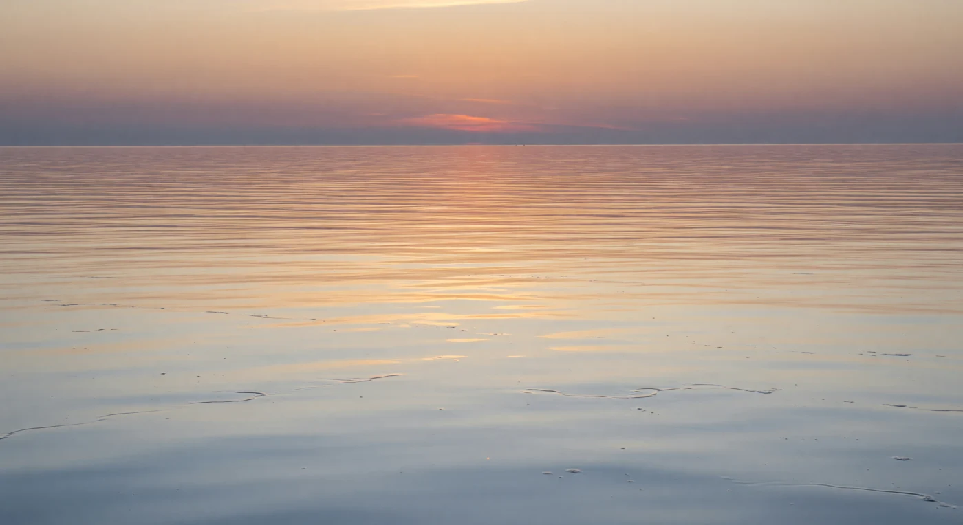 At the ocean's skin — a boundary measured not in fathoms but in micrometers — the air and sea negotiate their exchange across a film thinner than a human hair. Here, where wind stress has nearly vanished under a Beaufort 0 sky, surface tension governs everything: the sea-surface microlayer, a gel-rich lens of lipids, transparent exopolymers, and dissolved organics, stretches unbroken across the water, compressing the dusk light into long specular bands of copper and rose-gold that slide over undulations barely tall enough to cast a shadow. Beneath this luminous skin, the uppermost meters of the water column hold their own quiet world — neuston organisms, the larvae of countless pelagic species, and colonies of bacteria exploiting the microlayer's chemical richness, all suspended in a blue-gray transparency where the day's final photons still reach. The physics of gas exchange — carbon dioxide, oxygen, water vapor moving between ocean and atmosphere — slow almost to a standstill when wind speed drops this low, turning this glassy interface into a temporary seal between two vast reservoirs. Nothing breaks the stillness; the planet breathes in near-silence, the sea folding light back into sky as if the two have no clear boundary between them.