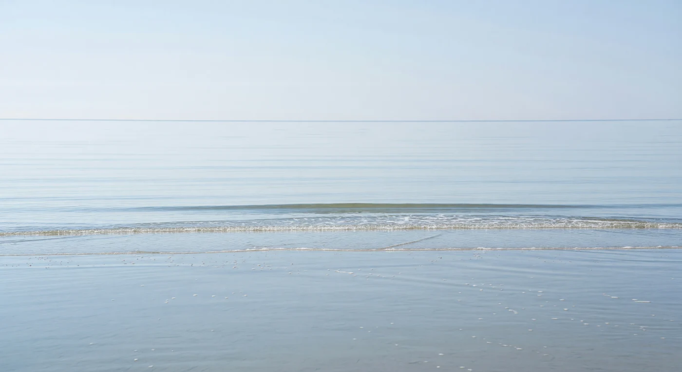 Op het grensvlak tussen oceaan en atmosfeer, waar wind en zee elkaar nauwelijks raken, regeert een wereld van uiterste stilte en subtiele fysica. Het wateroppervlak functioneert hier als een levende membraan — de zeemicrolayer, een film van amper enkele honderden micrometers dik, geconcentreerd met bacteriën, lipiden, polysacchariden en opgelost organisch materiaal dat zich opstapelt in de kalmte en een bijeenkomst vormt van het onzichtbare leven. Onder de dun trillende oppervlakte, waar causaal licht door helder water kaatst en de zandbodem van het ondiepe zandbank vaalbleek oplicht, fotosynthetiseren fytoplankton en produceren zij de zuurstof en biomassa die de gehele waterkolom voedt. De enkele langgerekte deining die het zandbank bereikt en breekt, geeft voor een fractie van een seconde uitdrukking aan de enorme energiereizen die golven afleggen — geboren in stormen op honderden kilometers afstand, eindigend hier in een fluisterlijn van wit schuim en fijn spuitzout. Wat van buiten als leegte lijkt, is in werkelijkheid een van de meest ecologisch actieve en klimatologisch bepalende lagen van de aarde, onophoudelijk gassen en warmte uitwisselend met de lucht erboven, in afwezigheid van elk oog dat het gadeslaat.