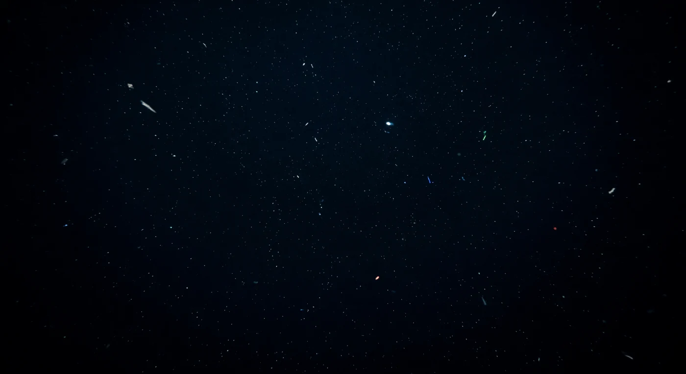 Os faróis do ROV projetam um cone estreito de luz fria que se dissolve em negro absoluto a poucos metros de distância, sem revelar fundo, parede ou qualquer forma viva — apenas o vazio ilimitado da coluna de água em todas as direções. Através desse feixe, uma chuva constante de neve marinha desce em diagonal: fragmentos translúcidos de matéria orgânica, exoesqueletos dissolvidos, fezes compactadas e muco bacteriano que afundam lentamente desde a zona fótica, centenas de metros acima, transportando carbono fixado pelo sol para um mundo que nunca o verá. A pressão aqui ultrapassa duzentas atmosferas, comprimindo o casco do veículo com uma força que tornaria qualquer erro fatal, e a temperatura da água paira próximo de dois graus Celsius, suficiente para abrandar o metabolismo de qualquer organismo que ouse habitar este espaço intersticial entre a superfície e o sedimento. Os LEDs de estado do veículo — âmbar, verde, vermelho escuro — piscam nas bordas do enquadramento como os únicos sinais de tecnologia num ambiente onde a bioluminescência seria a única outra fonte de fotões, caso algum animal passasse agora pelo campo de visão. A neve marinha continua a derivar, indiferente, conferindo a única sensação de escala e movimento neste vazio sem horizonte onde a distância se torna impossível de calcular.